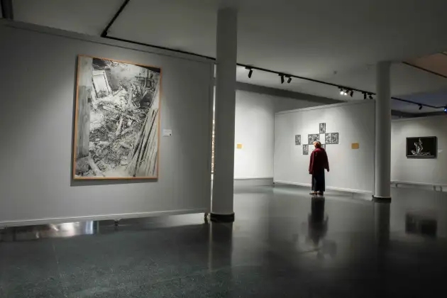 Fotografía de una sala de exposiciones en un museo, iluminada artificialmente y con paredes grises y suelo oscuro brillante. En primer plano, a la izquierda, se observa una gran obra enmarcada en blanco y negro sobre la pared con tema de escombros y tablones. Más al fondo, hay una persona de pie con chaqueta burdeos que contempla una agrupación de nueve dibujos enmarcados, dispuestos en forma de cruz en la pared central. Otra pared lateral muestra una pintura enmarcada con figura humana en blanco y negro. El espacio aparece segmentado por columnas blancas y transmite ambiente silencioso y contemplativo, propio de una exposición.