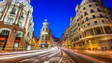 Fotografía nocturna de la Gran Vía de Madrid con cielo azul oscuro. La imagen se centra en edificios históricos iluminados a ambos lados de una avenida principal. Destacan los detalles arquitectónicos neoclásicos y barrocos en las fachadas, con columnas, balcones y esculturas. En el centro, la calle curva se llena de largas líneas de luces blancas y rojas, trazadas por el movimiento de los coches en larga exposición. No aparecen personas visibles. El ambiente transmite dinamismo urbano, con contraste entre la luz artificial de las farolas y la iluminación cálida de los edificios.