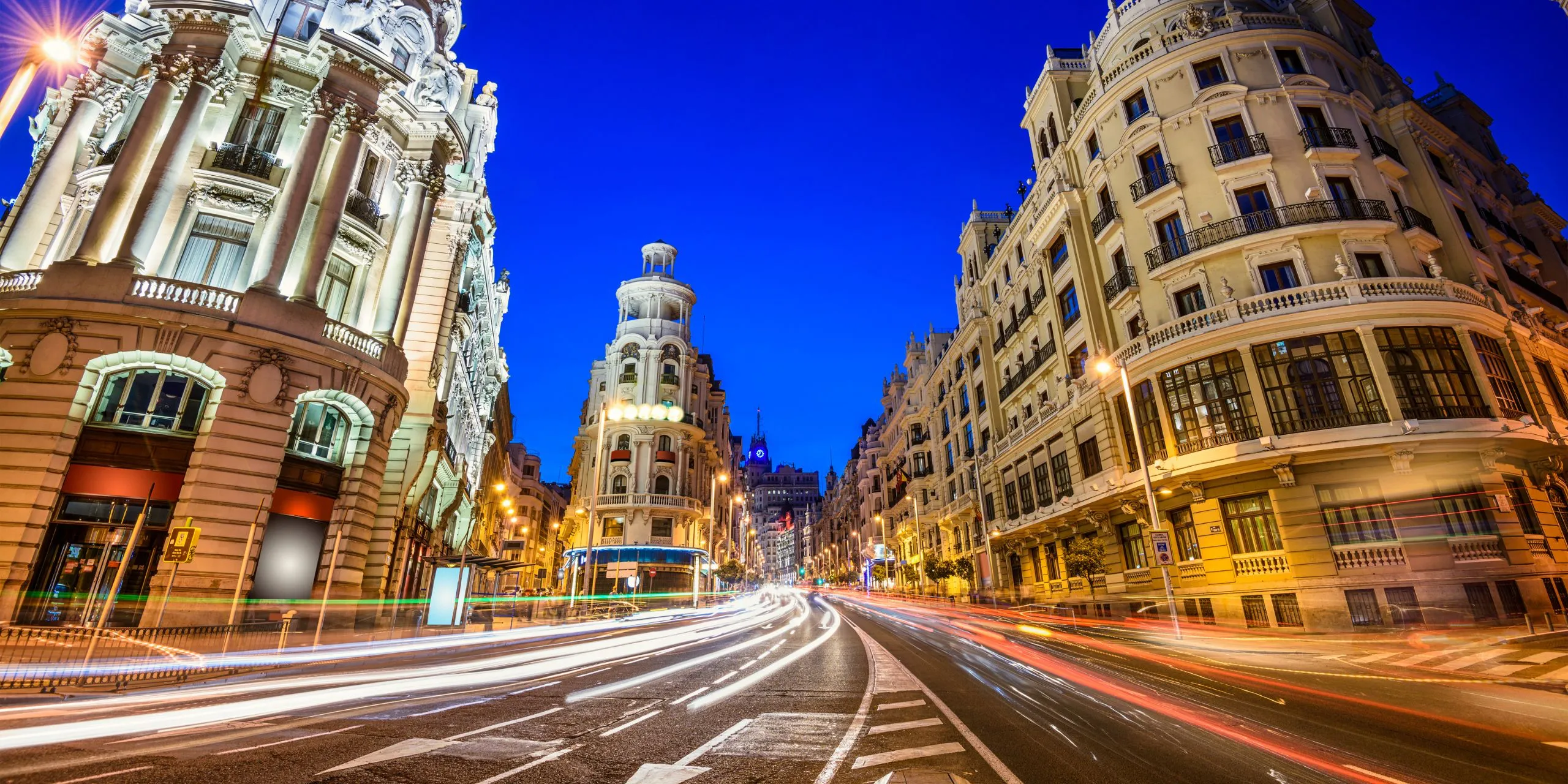Fotografía nocturna de la Gran Vía de Madrid con cielo azul oscuro. La imagen se centra en edificios históricos iluminados a ambos lados de una avenida principal. Destacan los detalles arquitectónicos neoclásicos y barrocos en las fachadas, con columnas, balcones y esculturas. En el centro, la calle curva se llena de largas líneas de luces blancas y rojas, trazadas por el movimiento de los coches en larga exposición. No aparecen personas visibles. El ambiente transmite dinamismo urbano, con contraste entre la luz artificial de las farolas y la iluminación cálida de los edificios.