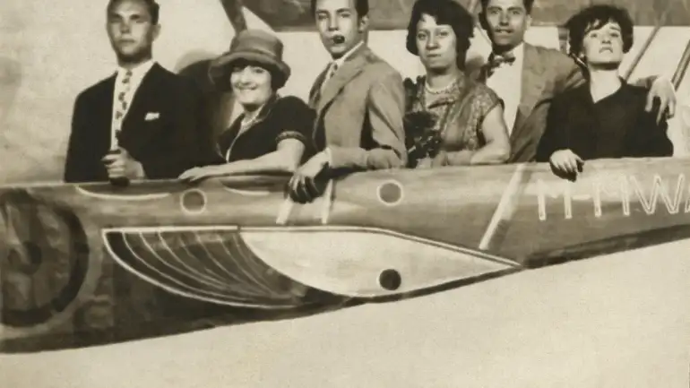 Maruja Mallo, Gregorio Prieto, Rosa Chacel, Concha Méndez y dos amigos en una verbena. Madrid, 1927 Museo Gregorio Prieto. Fotografía en blanco y negro tomada en Madrid en 1927 durante una verbena. Seis personas posan en línea dentro de un decorado con forma de avión, visible hasta la mitad inferior de sus cuerpos. De izquierda a derecha, se ve a un hombre joven de pie con expresión seria, vestido con traje oscuro y corbata clara con dibujos. Junto a él, una mujer sonriente con sombrero y vestido oscuro de bordes claros, identificada como Maruja Mallo. El siguiente es un hombre joven en traje claro que sostiene una pipa con la boca y mira a la cámara con gesto animado. A su lado, una mujer con vestido decorado y collar, identificada como Concha Méndez. Junto a ella, Gregorio Prieto, en camisa y pajarita, apoya su brazo izquierdo sobre el hombro de la mujer que está a su derecha. Esta última es Rosa Chacel, de cabello corto y oscuro, vestida de negro, con gesto sereno y la mano apoyada sobre el borde decorativo del supuesto avión. El fondo es una estructura pintada que simula una aeronave, con ventanas redondeadas y la matrícula “M-HW” visible en el lateral. La escena transmite el ambiente festivo propio de una celebración popular, típica del contexto urbano de los años 20.