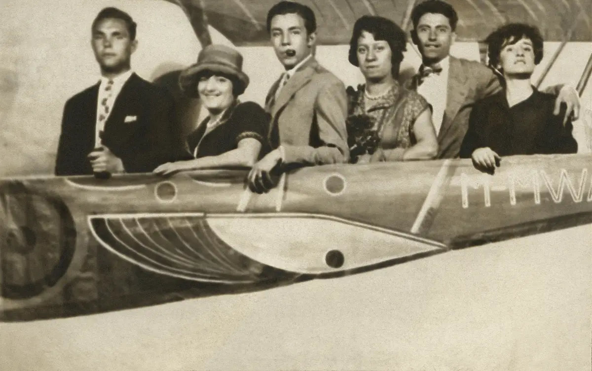 Maruja Mallo, Gregorio Prieto, Rosa Chacel, Concha Méndez y dos amigos en una verbena. Madrid, 1927 Museo Gregorio Prieto. Fotografía en blanco y negro tomada en Madrid en 1927 durante una verbena. Seis personas posan en línea dentro de un decorado con forma de avión, visible hasta la mitad inferior de sus cuerpos. De izquierda a derecha, se ve a un hombre joven de pie con expresión seria, vestido con traje oscuro y corbata clara con dibujos. Junto a él, una mujer sonriente con sombrero y vestido oscuro de bordes claros, identificada como Maruja Mallo. El siguiente es un hombre joven en traje claro que sostiene una pipa con la boca y mira a la cámara con gesto animado. A su lado, una mujer con vestido decorado y collar, identificada como Concha Méndez. Junto a ella, Gregorio Prieto, en camisa y pajarita, apoya su brazo izquierdo sobre el hombro de la mujer que está a su derecha. Esta última es Rosa Chacel, de cabello corto y oscuro, vestida de negro, con gesto sereno y la mano apoyada sobre el borde decorativo del supuesto avión. El fondo es una estructura pintada que simula una aeronave, con ventanas redondeadas y la matrícula “M-HW” visible en el lateral. La escena transmite el ambiente festivo propio de una celebración popular, típica del contexto urbano de los años 20.