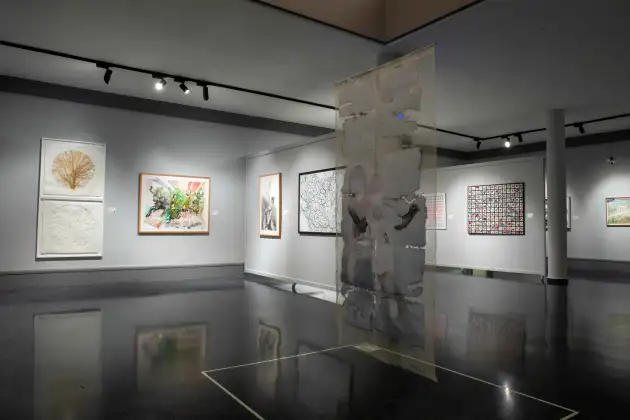 Sala de exposiciones temporales Museo Gregorio Prieto, vista de sala. Fotografía panorámica interior de una sala de museo contemporáneo, con paredes y columnas grises y suelo brillante. En la parte central destaca una gran obra vertical translúcida suspendida y enmarcada en un recuadro blanco sobre el suelo. El resto de paredes presenta obras de diferentes estilos: a la izquierda, un relieve vegetal y otro circular en blanco; una acuarela colorida; un dibujo realista en blanco y negro; obras abstractas de líneas negras o entramados de pequeños cuadrados rojos. Al fondo se ven más cuadros y diversidad de técnicas. La atmósfera es ordenada y sobria, con iluminación cuidada para resaltar cada obra, sin público presente en la imagen.