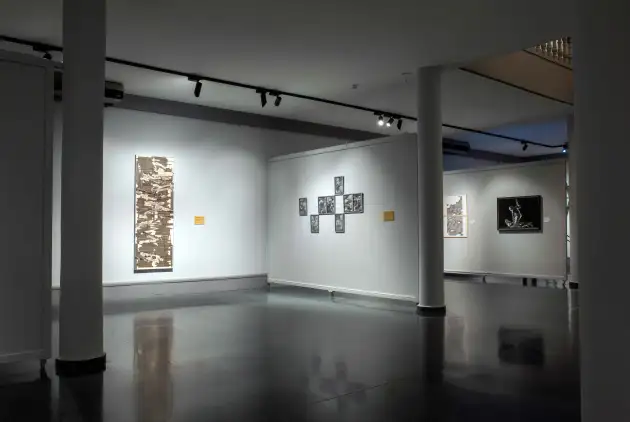 Sala de exposiciones temporales Museo Gregorio Prieto, obras ganadoras. Imagen de una amplia sala de exposiciones con paredes grises y suelo oscuro y brillante. Se aprecian varias columnas blancas repartidas por el espacio. En la pared izquierda cuelga una obra vertical alargada en tonos marrones y grises, iluminada por un foco. En la pared central, una agrupación simétrica de nueve dibujos enmarcados en blanco y negro forma una cruz, también muy iluminada. En la pared del fondo, otras dos obras: a la izquierda, un dibujo vertical, y a la derecha, una pintura en blanco y negro de una figura humana en pose dinámica. El ambiente es tranquilo, ordenado y de carácter museístico, sin personas a la vista.