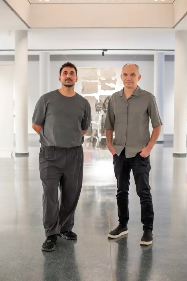 Fotografía vertical en el interior de una sala de exposiciones luminosa. Dos hombres adultos posan juntos en el centro de la imagen. El hombre de la izquierda, identificado como el artista Sebastián Bona, viste camiseta y pantalones oscuros y lleva el pelo corto; está de pie con las manos detrás de la espalda y cara neutra. A la derecha, José Miguel Chico López, con camisa gris claro, pantalón oscuro y zapatillas, está de pie con una mano en el bolsillo y la otra colgando, también con expresión neutra. El fondo revela paredes blancas, columnas y una obra abstracta de gran formato montada sobre paneles en la pared posterior, con zonas grises y trazos blancos, propia de una exposición artística. El suelo es brillante y oscuro, sin otros objetos ni personas visibles; todo el ambiente es ordenado y despejado.