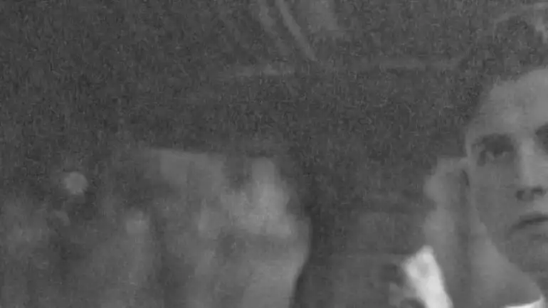 Retrato parcial de Gregorio Prieto en el Parque del Retiro, Madrid (1918–1919). La imagen en blanco y negro muestra un encuadre panorámico centrado en el rostro y busto de un joven Gregorio Prieto. Su expresión es seria y fija la mirada fuera del encuadre, hacia el espectador. Lleva una camisa clara abotonada hasta el cuello, sin chaqueta, y su cabello está cuidadosamente peinado hacia atrás con volumen. El fondo es difuso y está dominado por una gran fuente de piedra ornamentada, que se eleva justo detrás de él, parcialmente desenfocada, indicando que la escena transcurre al aire libre. La iluminación es tenue y natural, posiblemente capturada a la sombra de los árboles del Parque del Retiro en Madrid. El estilo de la fotografía, la ropa del retratado y la calidad de la imagen remiten a principios del siglo XX. Esta imagen corresponde a un fragmento de una fotografía más amplia realizada entre 1918 y 1919.