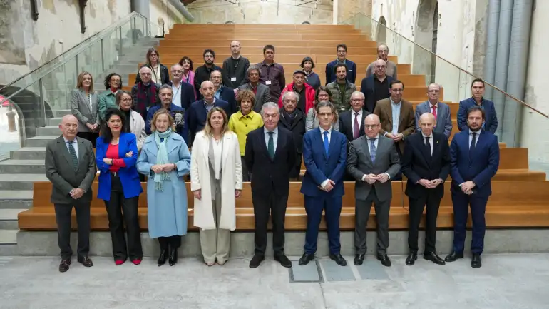 Fotografía en color, en formato horizontal, de un grupo numeroso de personas posando para una foto oficial en interior. El escenario es un gran espacio industrial rehabilitado: paredes altas con revocos desgastados y zonas de ladrillo visto, arcos abiertos en el lateral derecho, grandes conductos metálicos grises adosados a la pared y una cubierta elevada con estructura metálica y lucernarios que dejan entrar luz natural. En el centro del fondo hay una amplia grada de madera clara con peldaños largos; a ambos lados, barandillas de vidrio con pasamanos metálicos y escaleras de acceso. El grupo está distribuido en varias filas sobre los peldaños y el suelo. En la primera fila, situada en el nivel inferior, posan varias autoridades: en el centro se ve un hombre con traje oscuro y corbata verde; a su derecha y a su izquierda, otros hombres con trajes en tonos azul y gris, algunos con corbata. En el extremo izquierdo de esta fila aparece un hombre mayor con traje gris y corbata verde con lunares; cerca de él, una mujer con chaqueta azul intensa y blusa roja. También se distinguen dos mujeres con abrigos claros, una con abrigo azul claro y bufanda verde agua y otra con abrigo blanco, colocadas hacia el centro-izquierda. La mayoría de las personas de la primera fila miran a cámara con postura recta y manos delante del cuerpo o cruzadas. Detrás, sobre los peldaños de la grada, se agrupan los representantes de las fundaciones vinculadas a autores y artistas de la Generación del 27. Visten ropa de invierno (abrigos, chaquetas, bufandas) en colores sobrios —negros, grises, beiges y verdes— con algunas notas de color, como una chaqueta amarilla en el centro y una prenda roja en una de las filas intermedias. Varias personas llevan acreditaciones colgadas del cuello. Todas miran hacia la cámara, formando una composición simétrica, con la grada de madera como elemento central y el espacio industrial como telón de fondo.