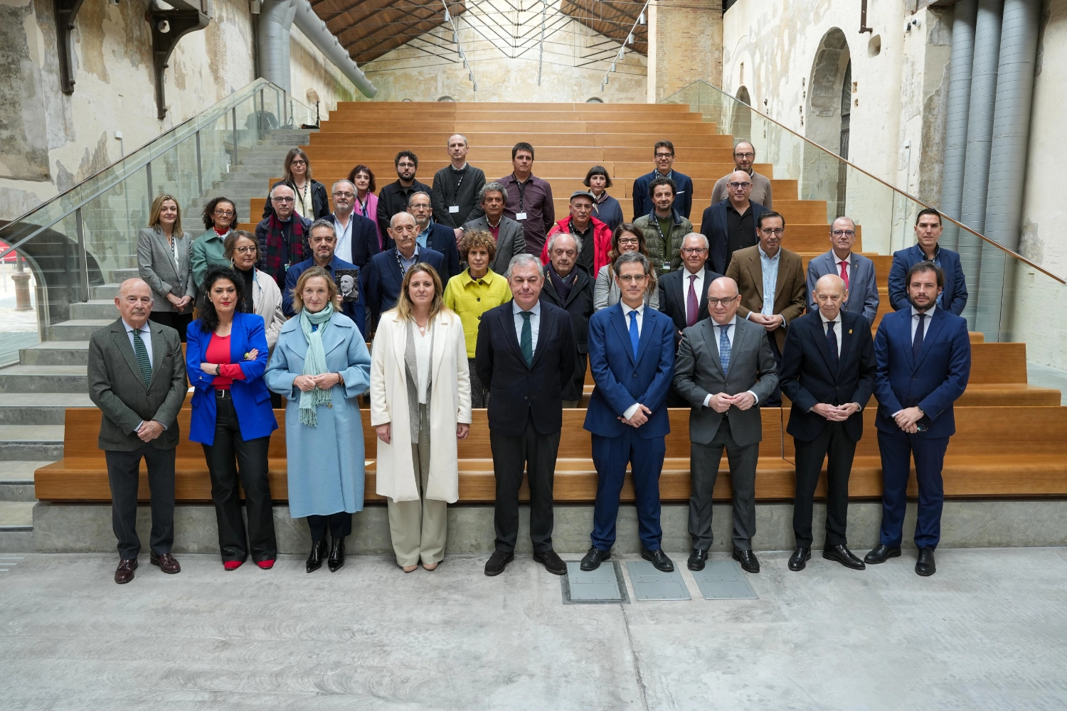 Fotografía en color, en formato horizontal, de un grupo numeroso de personas posando para una foto oficial en interior. El escenario es un gran espacio industrial rehabilitado: paredes altas con revocos desgastados y zonas de ladrillo visto, arcos abiertos en el lateral derecho, grandes conductos metálicos grises adosados a la pared y una cubierta elevada con estructura metálica y lucernarios que dejan entrar luz natural. En el centro del fondo hay una amplia grada de madera clara con peldaños largos; a ambos lados, barandillas de vidrio con pasamanos metálicos y escaleras de acceso. El grupo está distribuido en varias filas sobre los peldaños y el suelo. En la primera fila, situada en el nivel inferior, posan varias autoridades: en el centro se ve un hombre con traje oscuro y corbata verde; a su derecha y a su izquierda, otros hombres con trajes en tonos azul y gris, algunos con corbata. En el extremo izquierdo de esta fila aparece un hombre mayor con traje gris y corbata verde con lunares; cerca de él, una mujer con chaqueta azul intensa y blusa roja. También se distinguen dos mujeres con abrigos claros, una con abrigo azul claro y bufanda verde agua y otra con abrigo blanco, colocadas hacia el centro-izquierda. La mayoría de las personas de la primera fila miran a cámara con postura recta y manos delante del cuerpo o cruzadas. Detrás, sobre los peldaños de la grada, se agrupan los representantes de las fundaciones vinculadas a autores y artistas de la Generación del 27. Visten ropa de invierno (abrigos, chaquetas, bufandas) en colores sobrios —negros, grises, beiges y verdes— con algunas notas de color, como una chaqueta amarilla en el centro y una prenda roja en una de las filas intermedias. Varias personas llevan acreditaciones colgadas del cuello. Todas miran hacia la cámara, formando una composición simétrica, con la grada de madera como elemento central y el espacio industrial como telón de fondo.