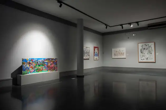 Espacio interior de exposición contemporánea, amplio y ordenado, con paredes grises y columnas blancas cilindricas. Se presenta una variedad de obras de arte enmarcadas, fundamentalmente dibujos y pinturas en técnica mixta con tonos vivos y pastel. En la pared de la izquierda, una obra compuesta por tres paneles de colores intensos (azul, verde, rojo, rosa) sobre una repisa blanca baja; el resto de las paredes tiene cuadros enmarcados con temática figurativa y abstracta, en distinto formato y estilo. La iluminación de focos ofrece luz directa sobre cada obra, resaltando los colores vibrantes y los detalles, sin presencia humana visible en la imagen. El ambiente es sobrio y propio de una inauguración museística.