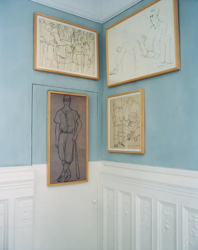 Rincón de Villa Josie con cuatro grabados originales de Gregorio Prieto enmarcados y colgados en la esquina de una pared azul claro y blanca, sobre un zócalo decorativo clásico. Los grabados, realizados en técnica de línea negra sobre papel claro, representan figuras humanas y deportivas en distintas posturas: en la parte superior izquierda, un grupo de jugadores en actitud deportiva; arriba a la derecha, figura masculina sentada en un sillón; en el centro, vertical, jugador de pie con gorra y bastón; a la derecha, dibujo de dos figuras masculinas sentadas, con piernas y brazos cruzados. La estancia es luminosa y serena, con paredes lisas celestes y moldura blanca en la parte inferior. Créditos de la imagen: Pablo Zamora para la Revista AD Best of España.